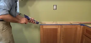 A person applying adhesive with a caulk gun to the top edge of wooden cabinets in preparation for countertop installation.