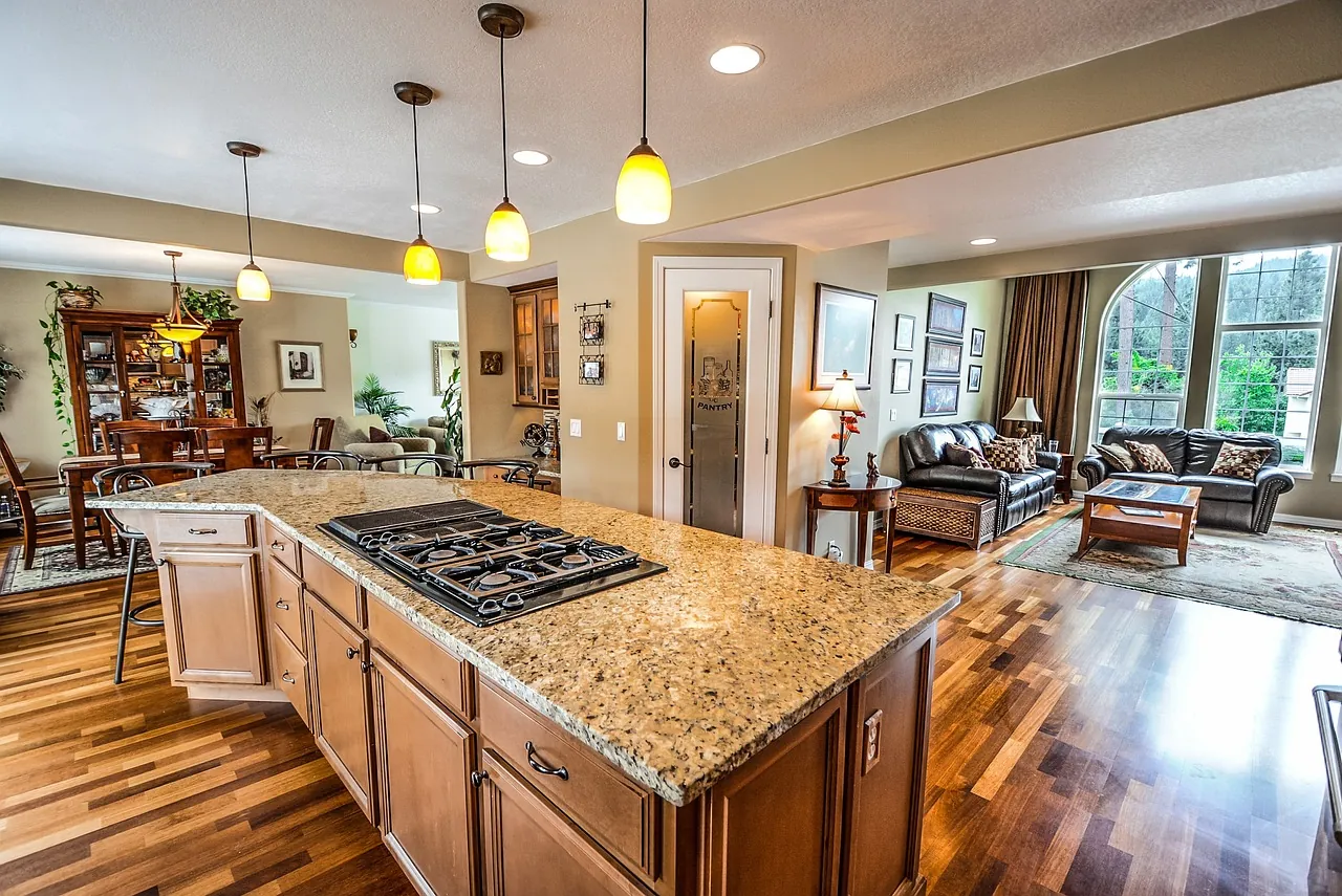 Open-concept kitchen with a large granite countertop island, wood cabinetry, pendant lighting, and a view into a cozy living and dining area with natural light streaming through large windows.